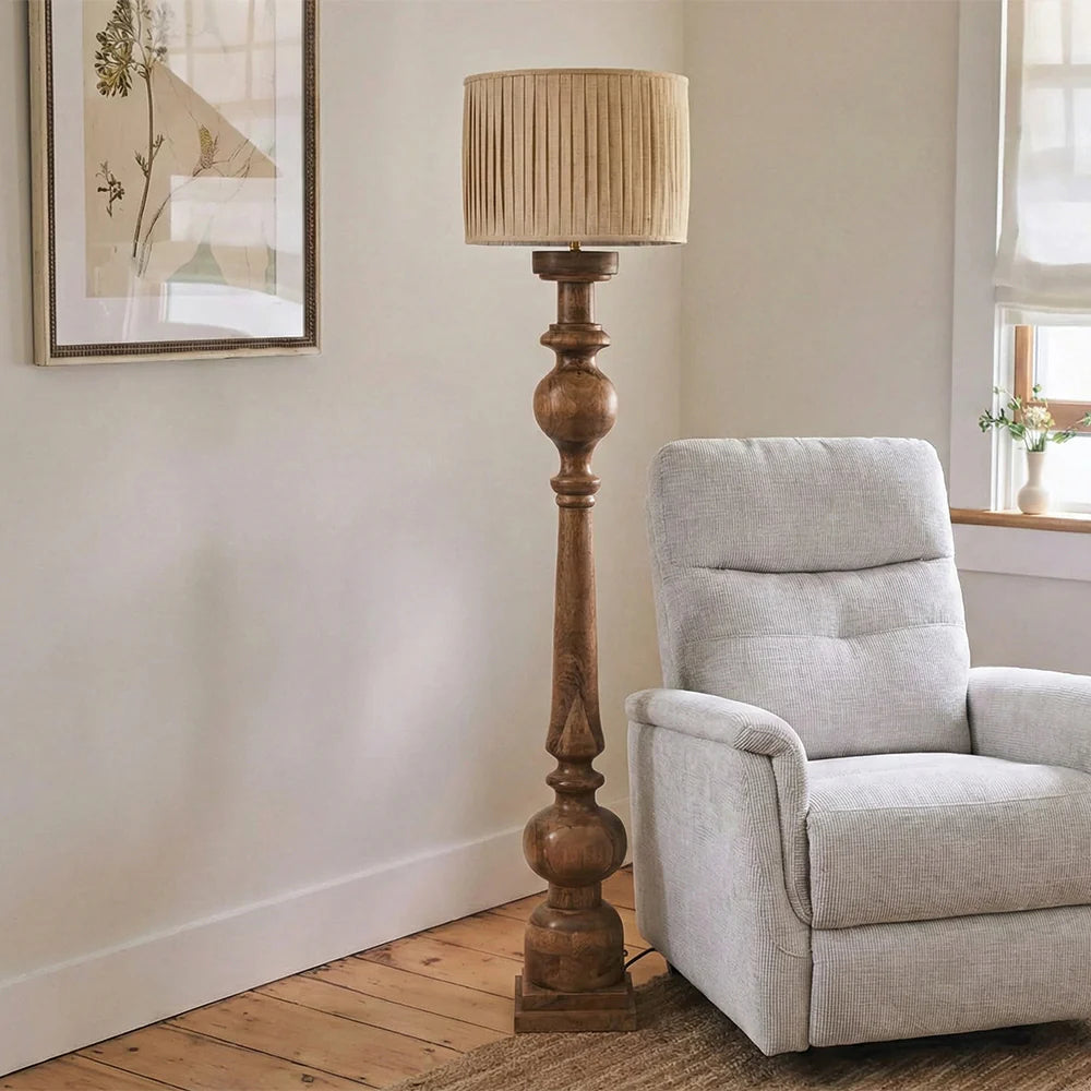 Wooden floor lamp with pleated shade next to a gray armchair in a room with a painting on the wall.