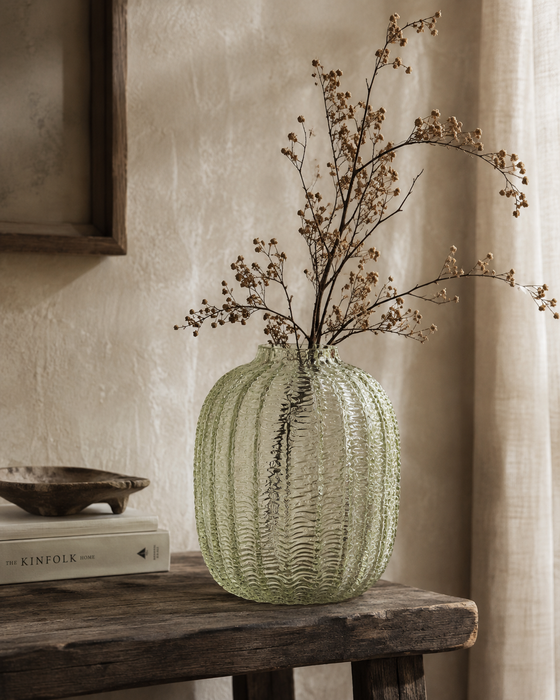 A ribbed, textured light green glass vase filled with dried baby's breath stems, displayed on a rustic wooden table next to a book in a warm, neutral-toned room.