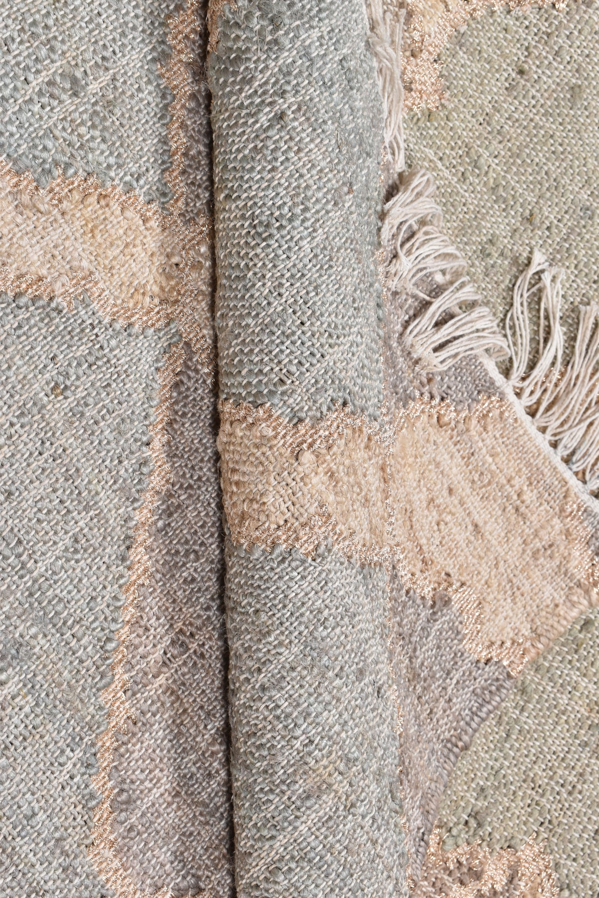 A close-up of a woven textile with a folded section showing fringed edges, in muted gray, beige, and soft pink tones.