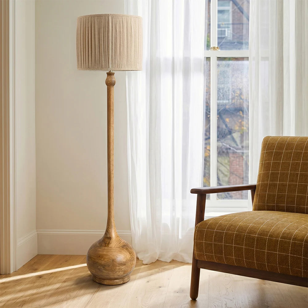 Wooden floor lamp with a beige shade next to a brown checkered armchair in a room with white curtains.