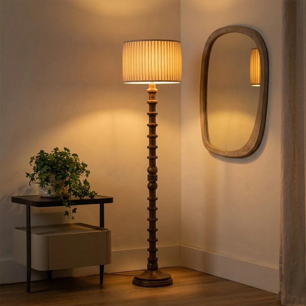 Wooden floor lamp with a textured shade next to a mirror on a wall, in a softly lit room.