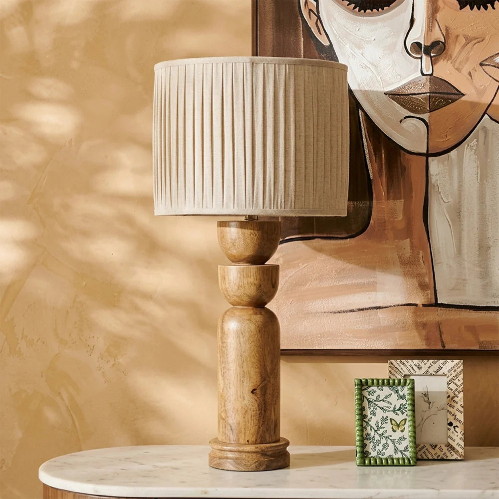 Wooden table lamp with pleated beige shade on a marble surface, with decorative items and a textured wall in the background.